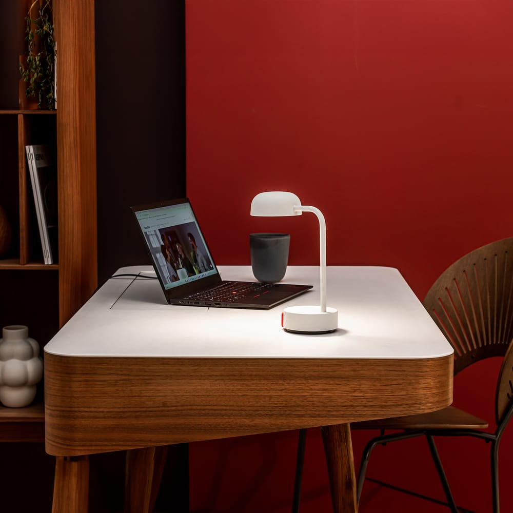 A modern white desk lamp with a curved neck and round head providing task lighting on a wooden table, with a laptop, cup, and decorative items in a room with a red wall.