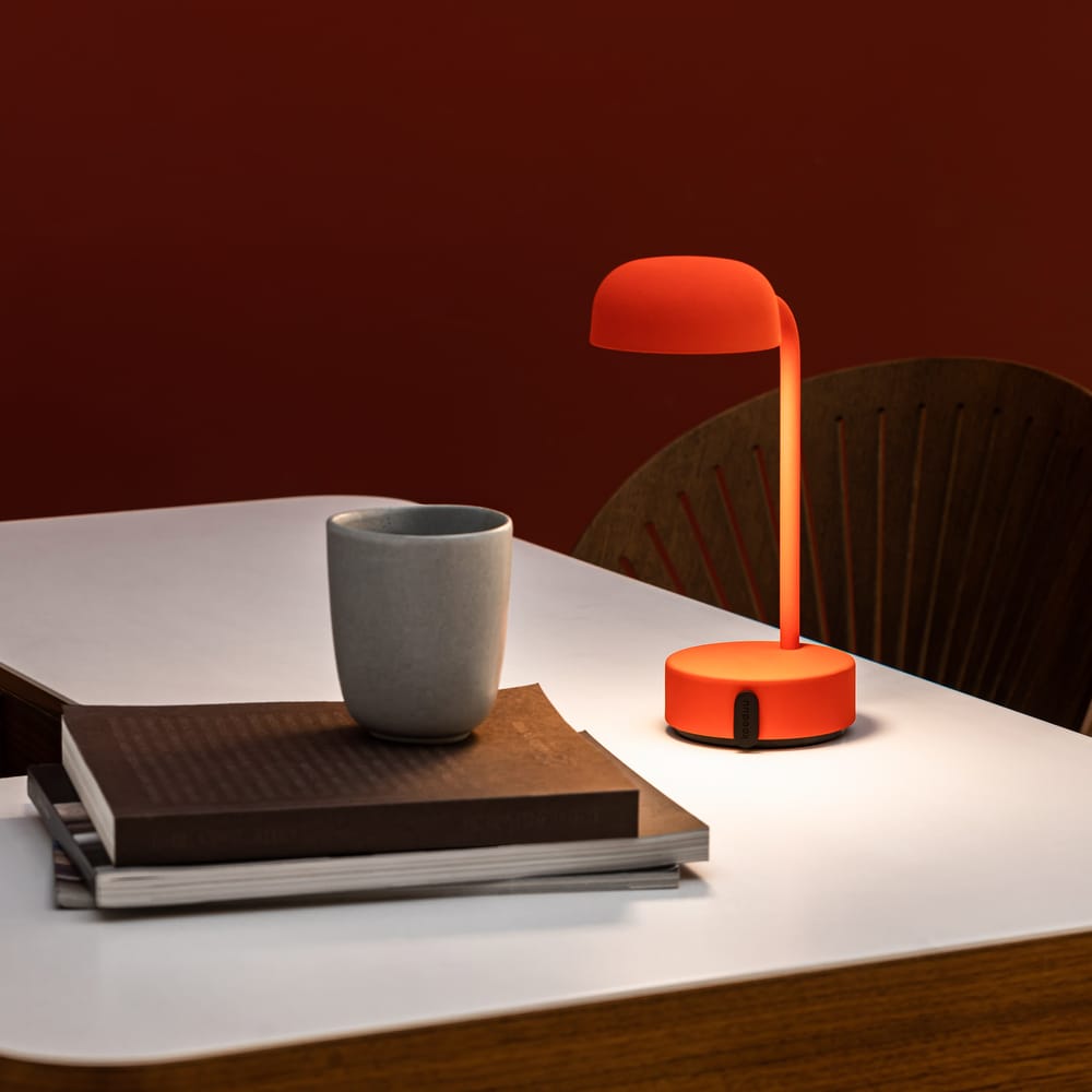 A vibrant orange desk lamp stands on a white table against a red wall, next to a gray mug and a stack of brown hardcover books, adding a pop of color to a modern workspace.