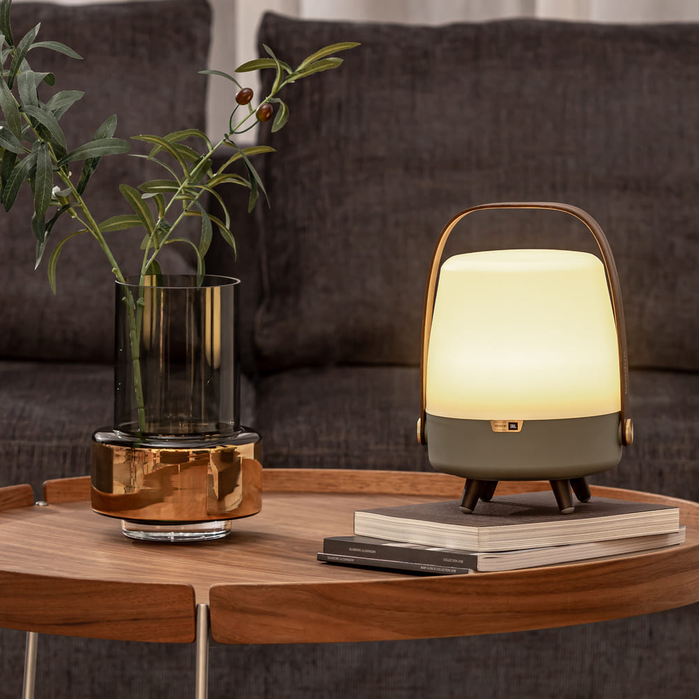 A stylish LED speaker lamp with a warm, ambient glow, featuring a golden handle, sits on a wooden table beside a stack of books and a clear vase with greenery, against a dark textured sofa backdrop.
