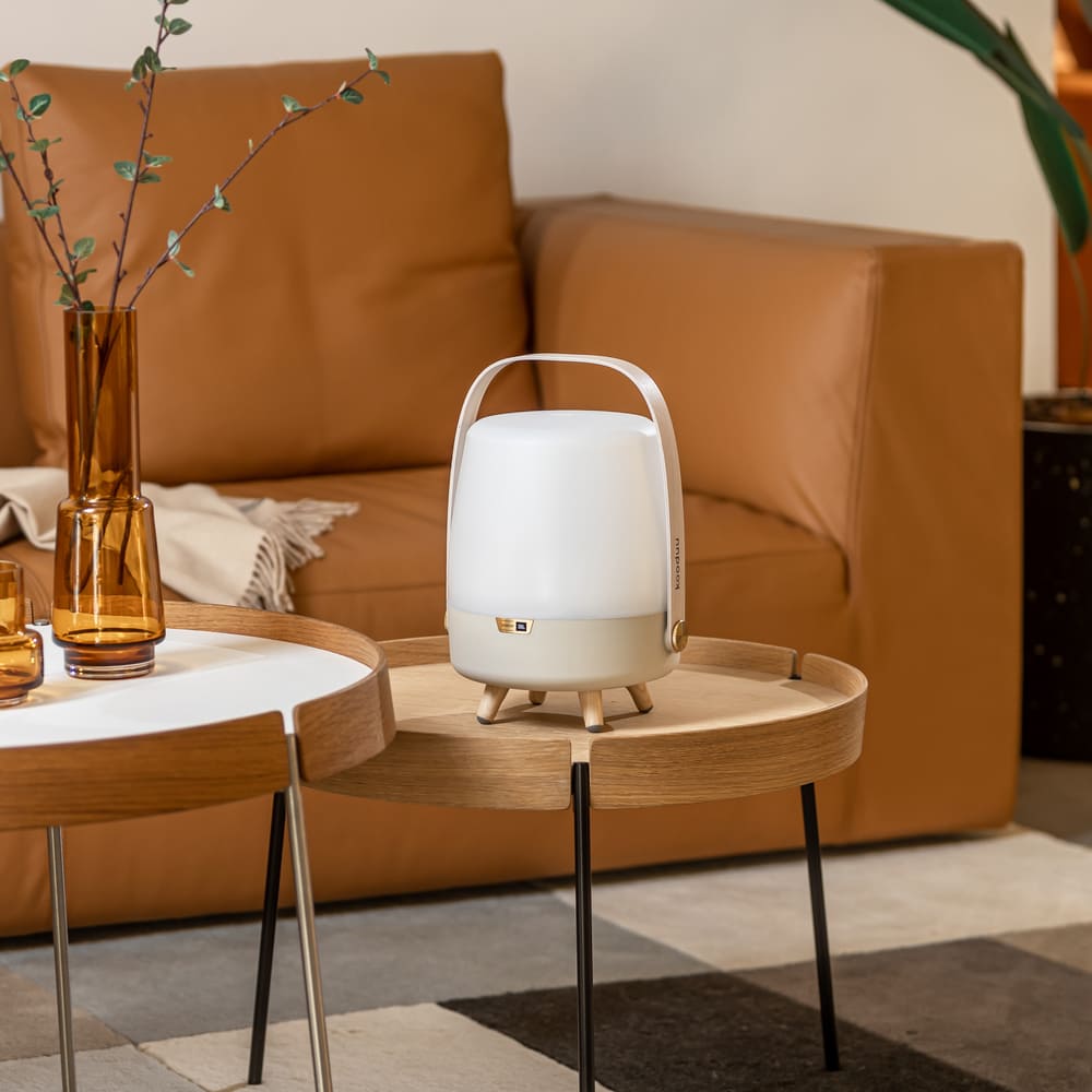 A modern white LED speaker lamp with a chic ivory handle, positioned on a round wooden coffee table near an amber glass vase with sprigs, complementing a tan leather couch in a cozy living room setting.