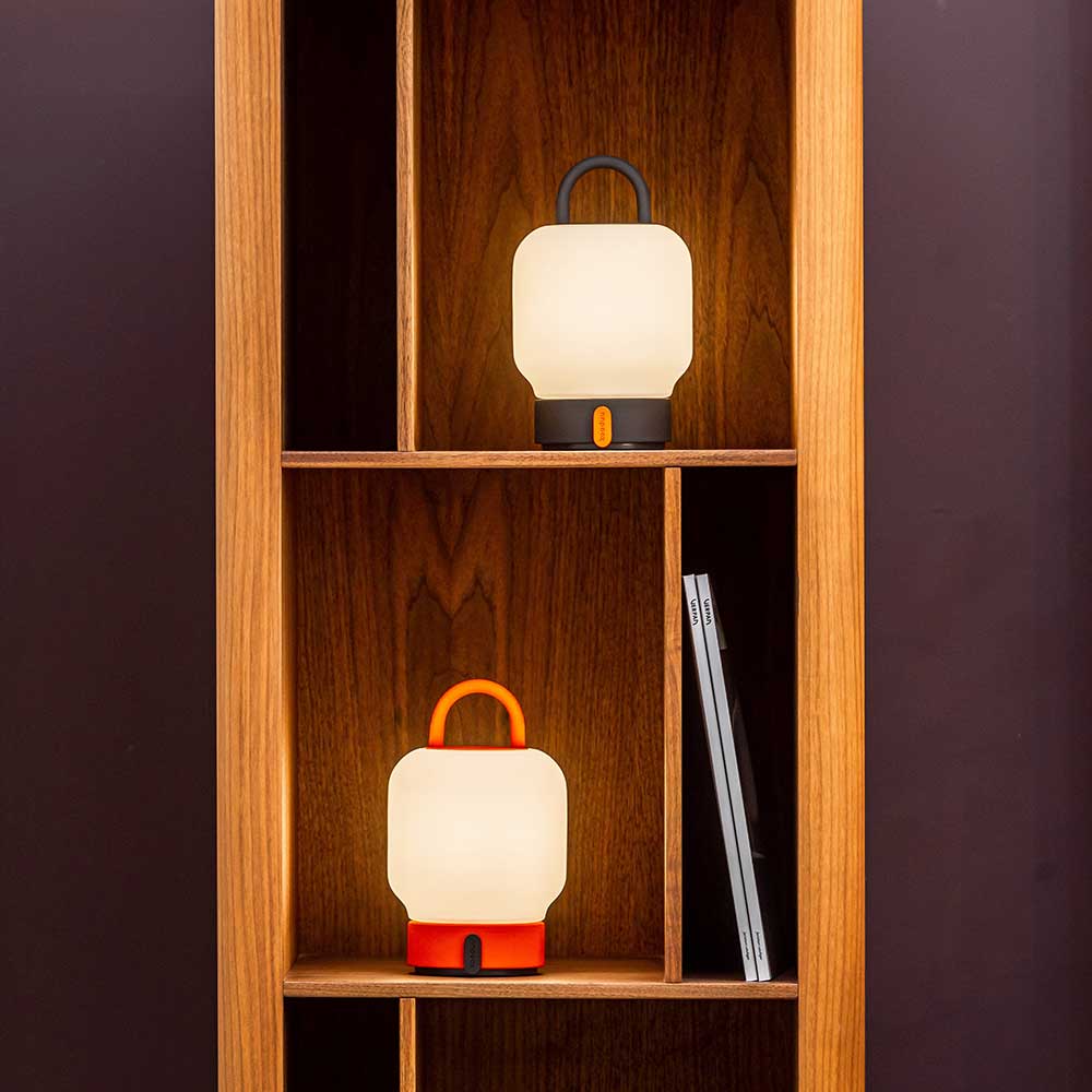 Two LED lantern table lamps on wooden shelves, one with an anthracite handle on the top shelf and one with an orange handle on the bottom shelf, with a magazine placed beside the lower lamp.