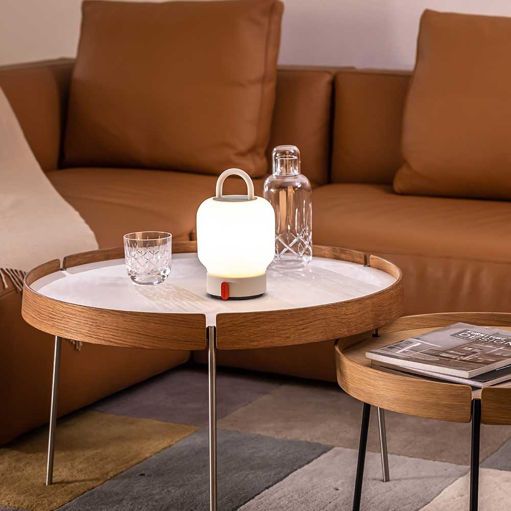Rechargeable LED table lamp with carrying handle on a round wooden side table, accompanied by a crystal glass and a clear water carafe, against the backdrop of a brown sofa with cushions.