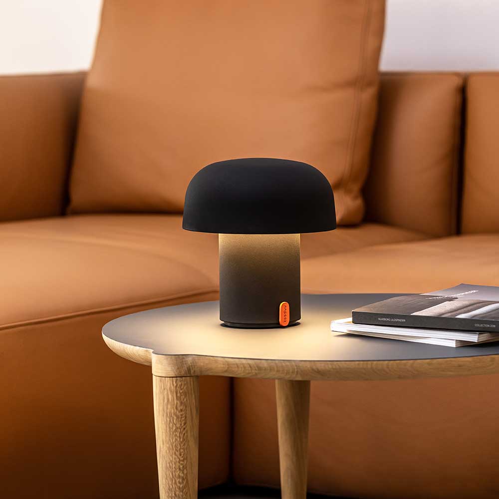 A contemporary anthracite-colored LED table lamp with a mushroom-like shape, placed on a round wooden side table next to a stack of books, against a backdrop of a tan leather sofa.
