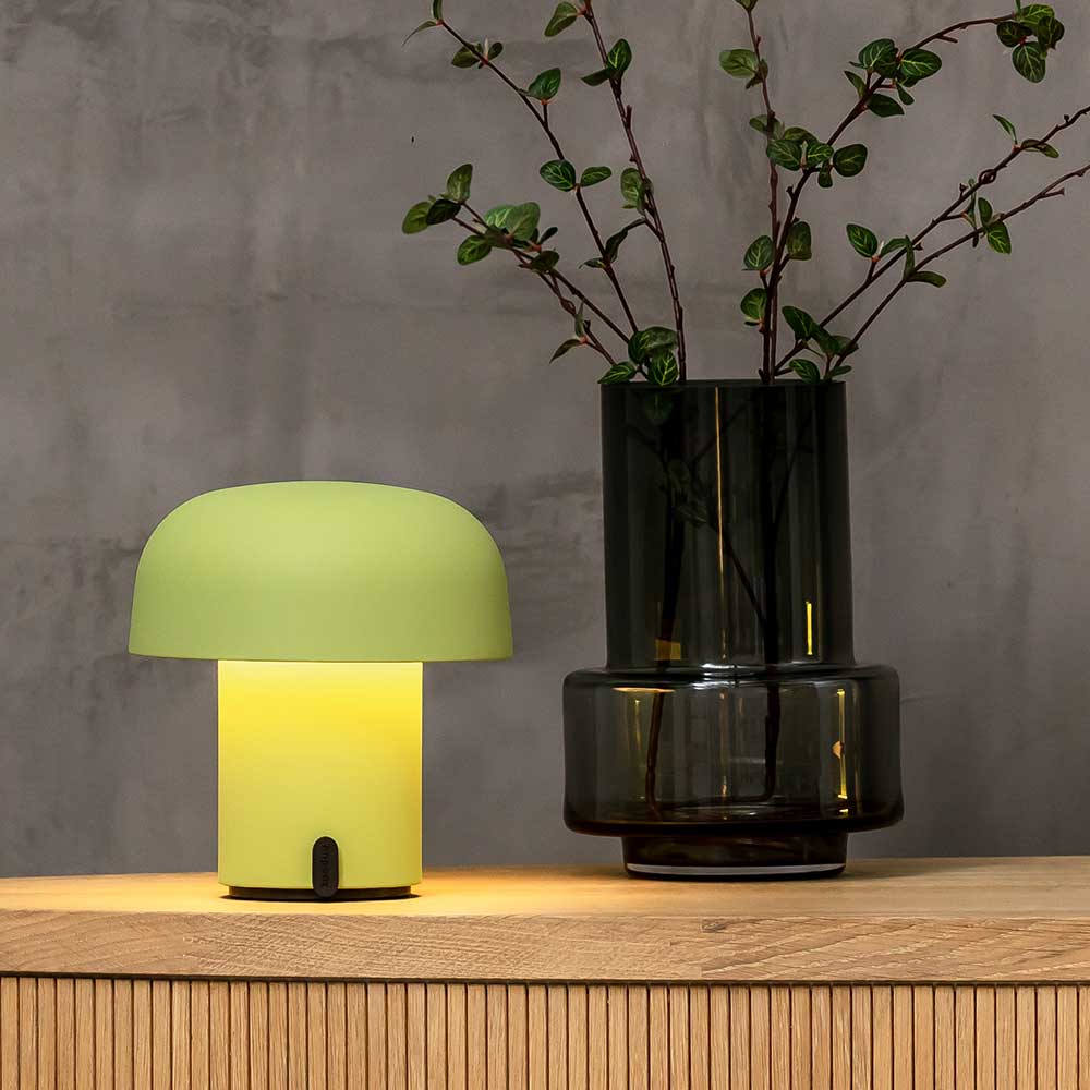A modern LED table lamp with a moss green shade glowing warmly, situated on a wooden surface next to a dark glass vase with sprouting green branches, against a grey concrete wall.