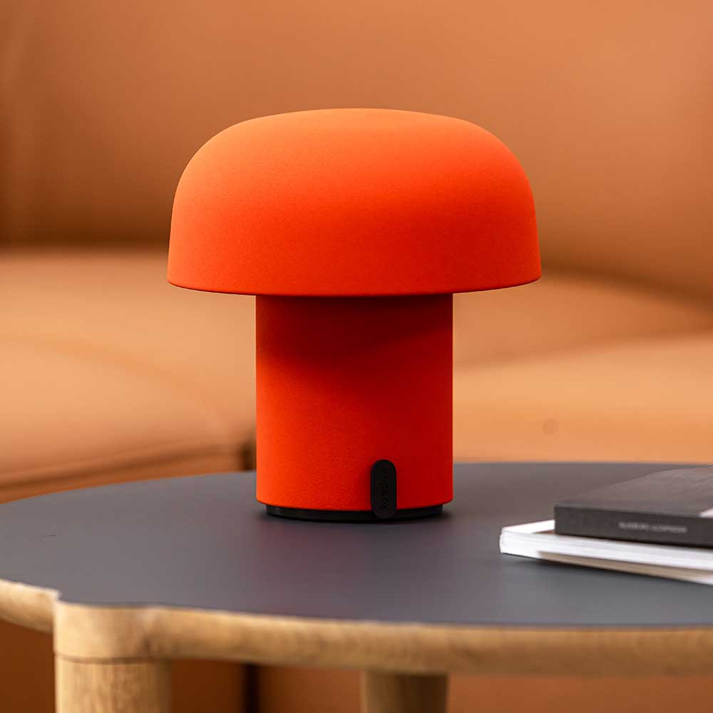 An orange LED table lamp with a contemporary design on a dark grey tabletop, featuring a soft, matte finish, placed beside a black hardcover book, with a warm tan sofa in the background.
